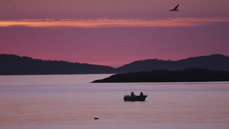 a small fishing boat carrying two fishermen floats peacefully on calm water during sunset, with silhouetted islands in the background.