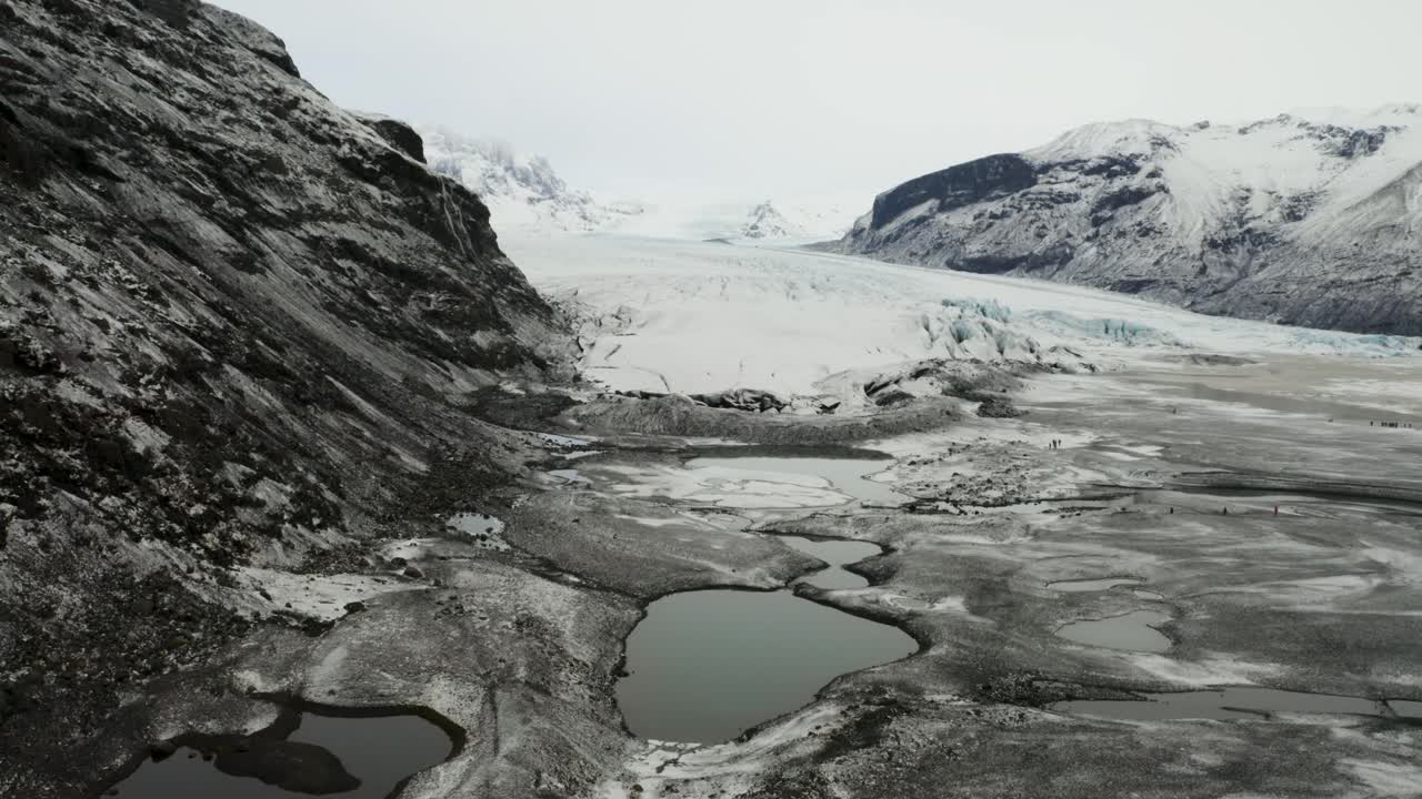 Captivating drone footage revealing the jagged crevasses of the glacier as it flows into a muddy meltwater lagoon surrounded by rugged cliffs.