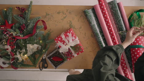 a top-down plan. fully visible the table with the decorations. female hands put and finalize christmas gift wrapped in craftool paper on a wooden table. bandaging tape