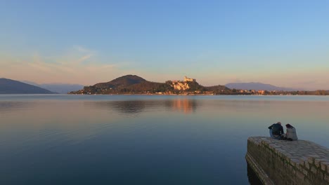 Romantisches-Paar-Sitzt-Am-Steg-über-Dem-Lago-Maggiore-Und-Blickt-Bei-Sonnenuntergang-Auf-Das-Schloss-Angera-Im-Hintergrund,-Italien