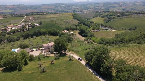 Wide-angle-drone-shot-following-a-car-driving-to-a-remote-village-in-the-countryside-of-Italy-surrounded-by-olive-trees-and-a-mountain-in-the-background-in-4k