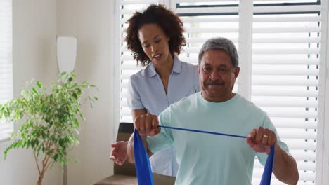 Mixed-race-female-physiotherapist-helping-senior-man-exercise-using-exercise-band