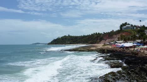 dolly in aerial drone shot of the tropical tibau do sul peninsula passing waves crashing into algae covered rocks and approaching the giz beach surrounded by cliffs in rio grande do norte, brazil