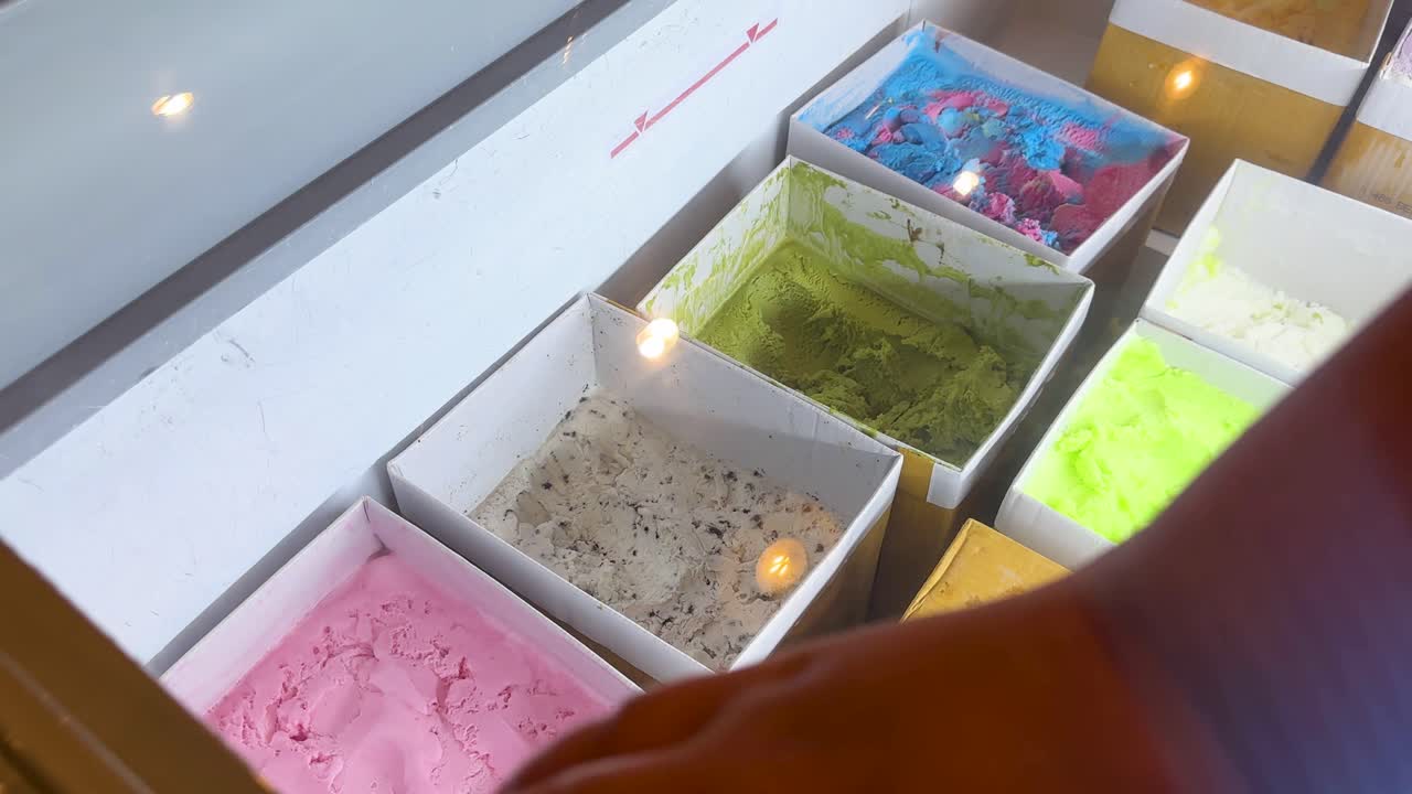 A hand chooses from colorful ice cream tubs in a well-lit display at a Bangkok market