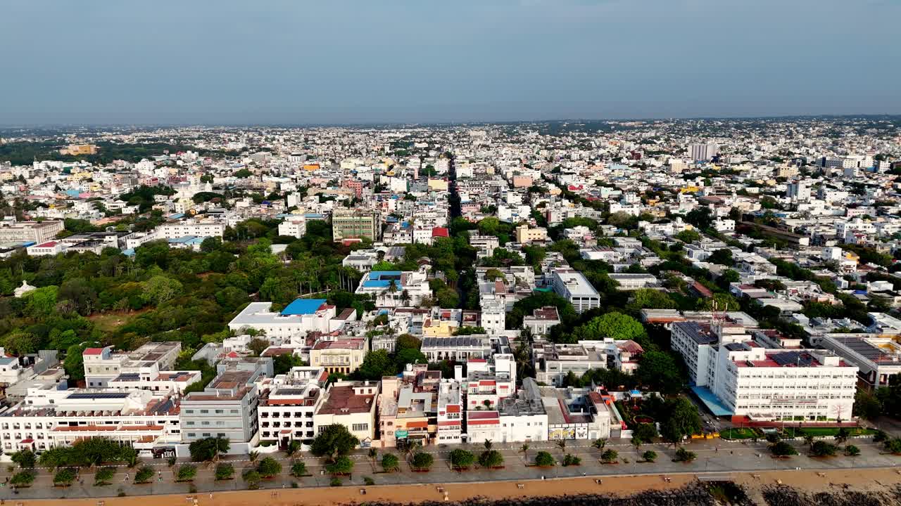 Aerial footage of White Town, Puducherry, a densely populated city with a prominent central road. The cityscape is a mix of buildings and lush green trees, showcasing a blend of urban life and nature