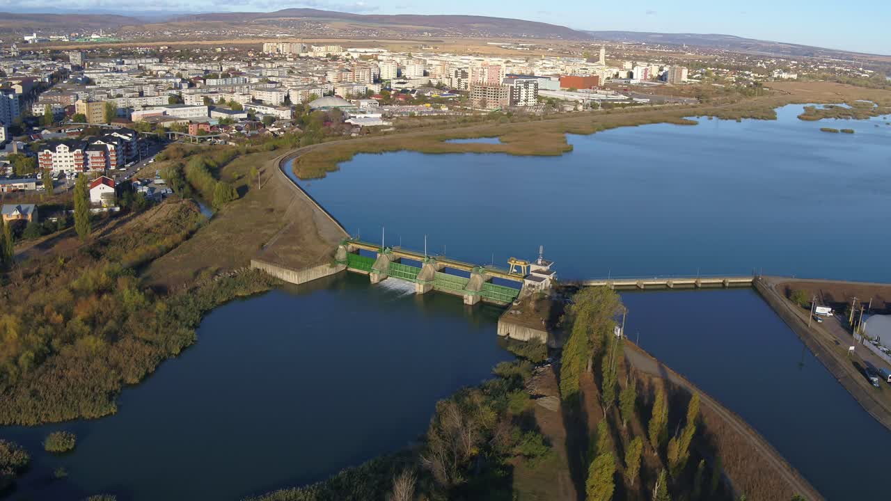A cinematic aerial shot slowly pushes in towards a hydroelectric dam and its reservoir. The city of Bacau is visible in the background on a sunny autumn afternoon