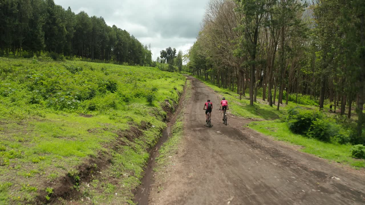 toma aérea de drones volando sobre atletas de maratón en bicicletas de montaña en bicicleta por un camino de tierra a través de un campo de hierba y tierras de cultivo entrenamiento muy pesado en el bosque de la selva tanzania después de la toma 4k