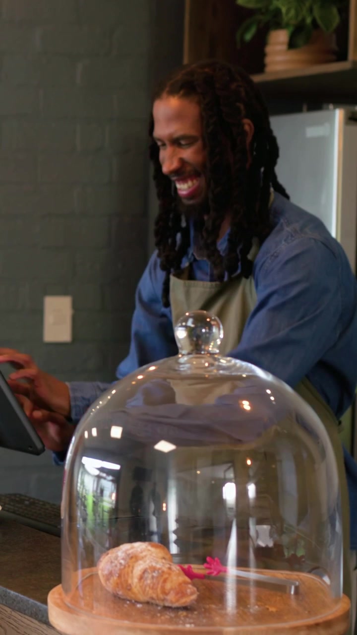 Vertical video: Hearing guest calling barista tapping POS reaching pastry dome at cafe green apron