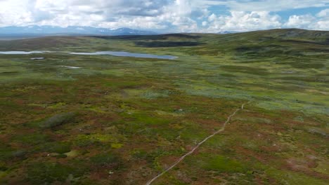 Gorgeous backward flying and revealing aerial drone shot of two hikers walking on a lonely narrow mud road in Sweden grassy green and brown landscapes with hills and mountains visible in the horizon.