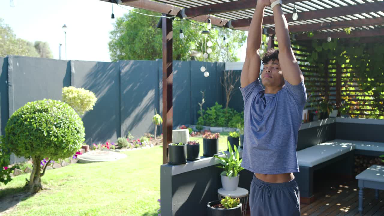 African American man in tee standing on deck under pergola inhaling, stretching arms toward lights