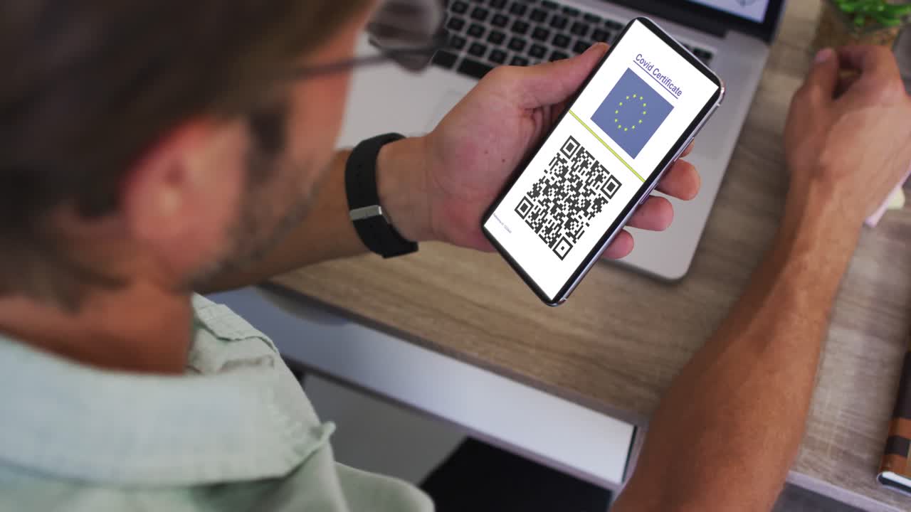 Man at desk holding smartphone with covid vaccination certificate, eu flag and qr code on screen