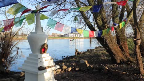 pequeño adorno de estupa budista en el borde del río ijssel en zutphen rodeado de árboles y vegetación con coloridas banderas de oración ondeando en el viento y detrás del barrio de noorderhaven