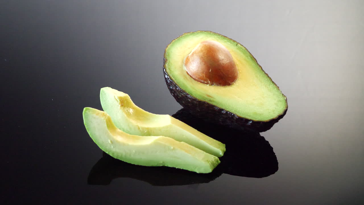Un avocado maturo tagliato a metà con il nocciolo e due fette pronte isolate su uno sfondo a specchio nero