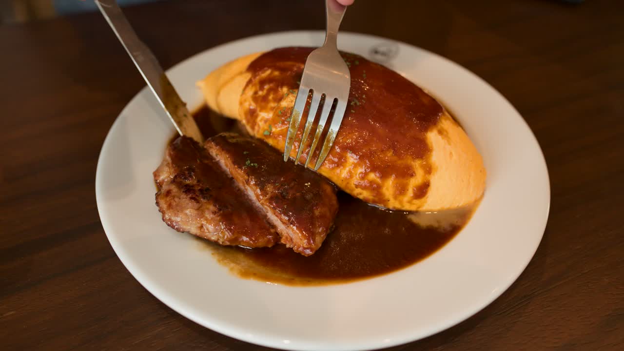 Hand slices juicy hamburg steak next to omelette rice, brown sauce, under warm restaurant lighting