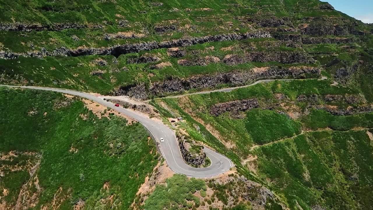 Aerial view of winding road at Miradouro Lombo do Mouro, Madeira, surrounded by lush green cliffs and dramatic terraced hillsides under clear blue sky, showing natural beauty and engineering marvels