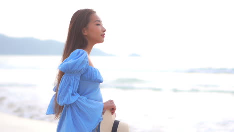 Slow-motion-view-of-beautiful-Asian-woman-standing-on-island-beach-in-blue-dress-holding-hat-looking-out-to-sea