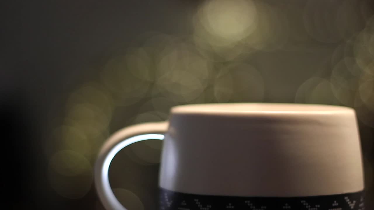 Steaming hot coffee in a beautiful mug in indoor setting with bokeh of lights in background. Slow sideways camera movement. Cup of tea captured in Full HD 1080p handheld shot.