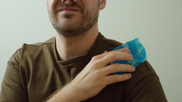 Close-up of a man applying a cold pack to his shoulder for cooling relief. Concept of muscle soreness, recovery, home therapy, and physical discomfort treatment