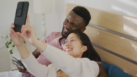 happy diverse couple taking selfie and lying in bedroom