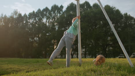trainer with hands gripping rod while stretching body on sunny green field outdoors as dog sniffs ground nearby in peaceful park environment surrounded by trees and natural light under clear sky