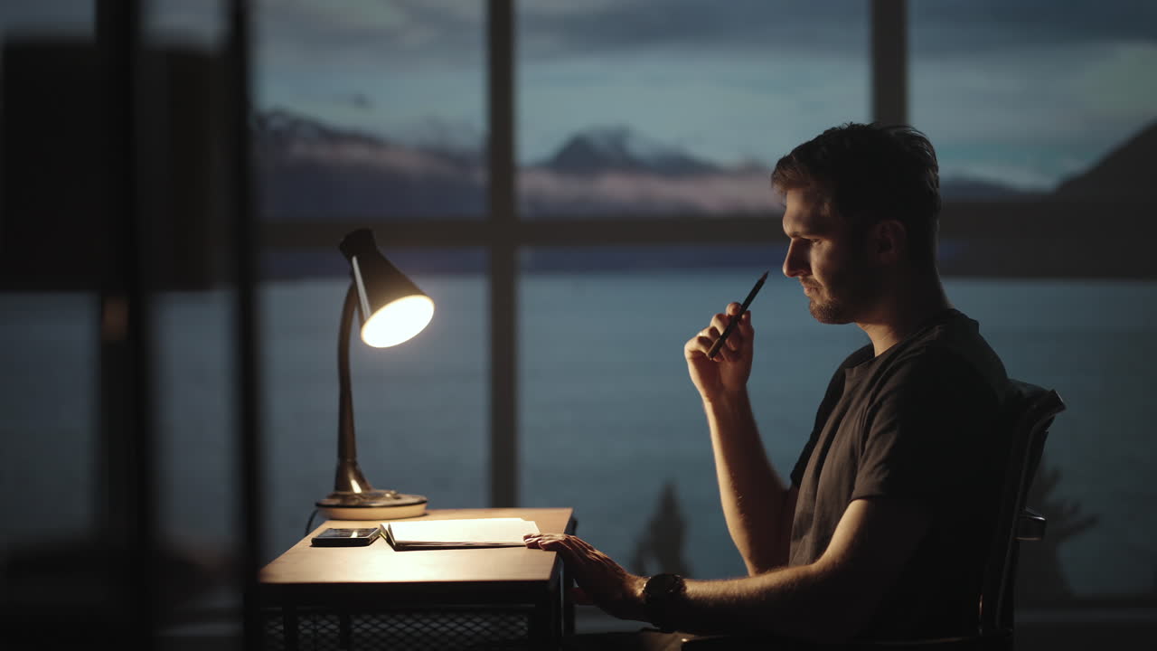 A thoughtful man sitting at a table with a desk lamp writes with a pen. Dark time of the day twilight on the background of the window a sad man thinks and writes