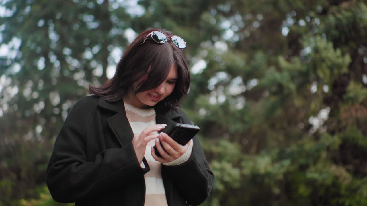 부드러운 보케 나뭇잎 아래 수목원에 서서 휴대폰 두 대 화면을 밀고, 캐주얼 코트를 입고, 머리에 선글라스를 얹고, 집중한 표정으로 차분한 야외 분위기를 내는 우아한 여성