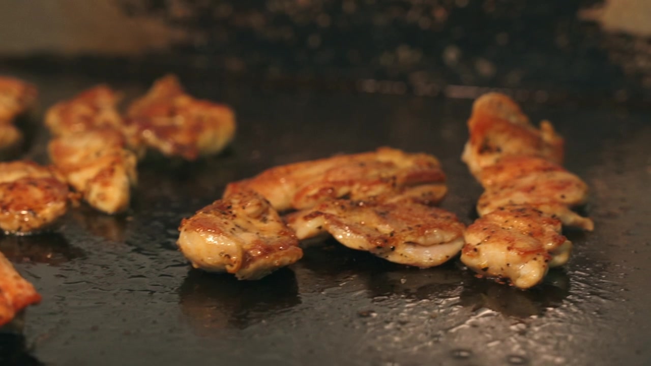 Inquadratura statica ravvicinata di pezzi di pollo marinato che cuociono e sfrigolano su una piastra commerciale rovente.