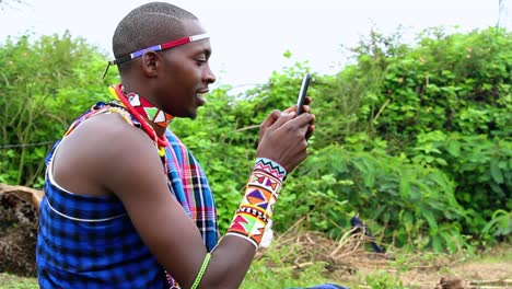 young moran male texting with a phone, sitting outdoors in forests of africa