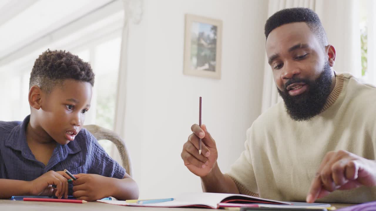 Happy african american father and son sitting at table and doing homework together, in slow motion