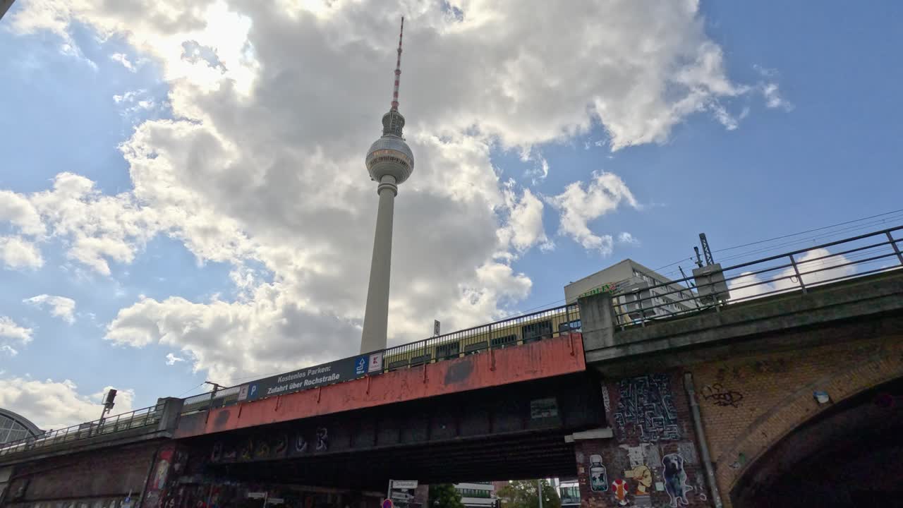 A red commuter train crosses a graffiti-covered bridge beneath the Berlin TV Tower, with dramatic clouds and sunlight overhead. Slight camera tilt adds dynamic movement