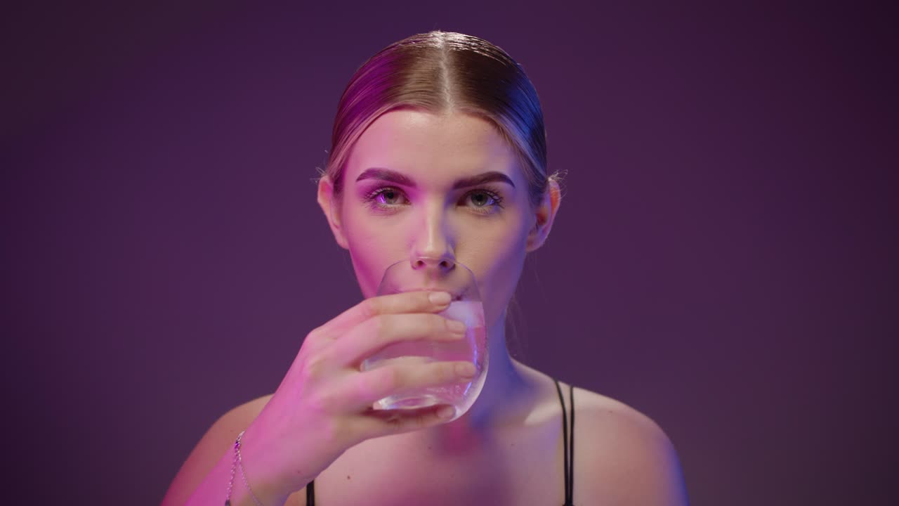 Splendida donna caucasica beve acqua da un bicchiere trasparente, sfondo scuro.