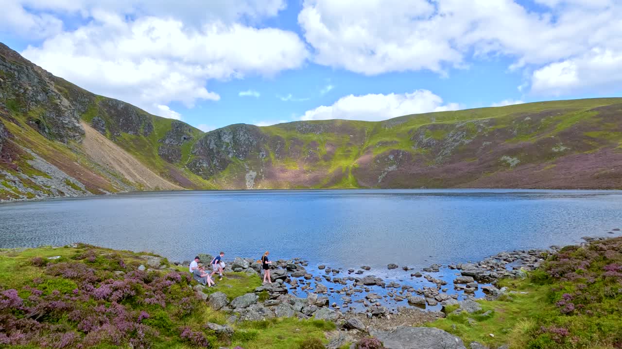 A group of hikers stands near the rocky shore of Loch Brandy, surrounded by heather and dramatic hills under bright daylight with a wide, steady camera view
