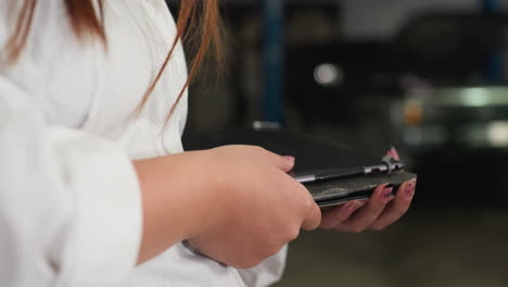 close up of woman in white lab coat using tablet in automotive workshop, recording data or analyzing information, focus on hands interacting with digital device in industrial setting