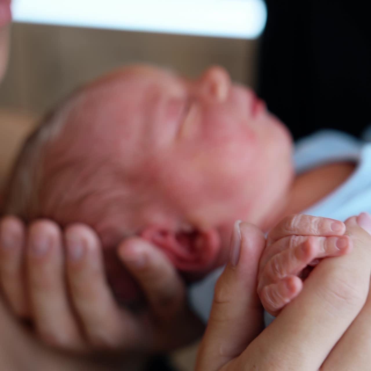 Tiny newborn child wearing blue romper in the parent's hands. Loving mother is holding cute fingers and kissing baby on the head. Close up