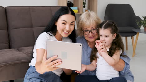 grandmother, mother and little girl having video call via tablet sitting on floor at home