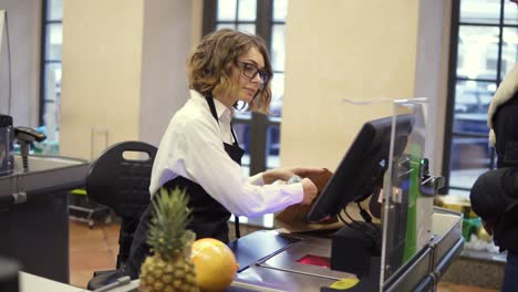 Cheerful-slender-saleswoman-in-white-shirt-and-black-apron-scanning-product,-fruits-at-checkout-counter-in-bright-supermarket-and-putting-it-into-brown-paper-bag-while-unrecognizable-customer-is-waiting