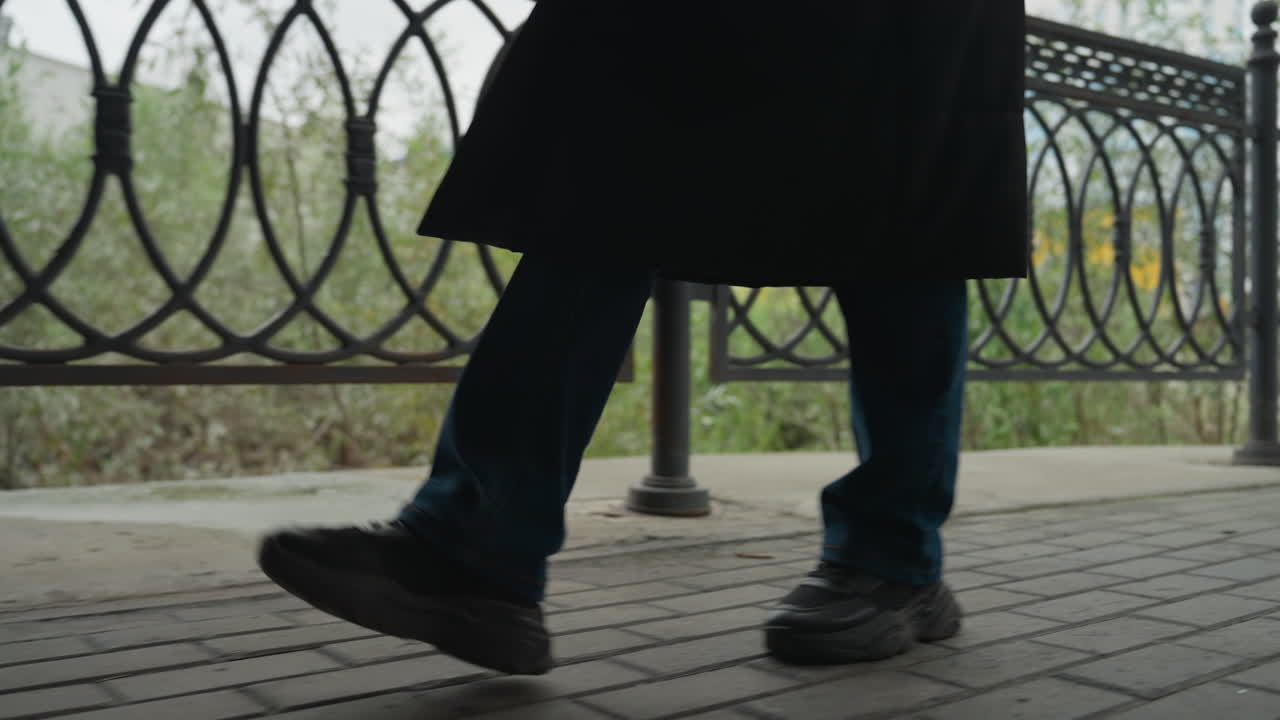 person walking along iron railing, jeans and coat visible, closeup of legs and shoes on wet cobblestone sidewalk, steady stride conveying solitary urban commute, muted overcast light and damp