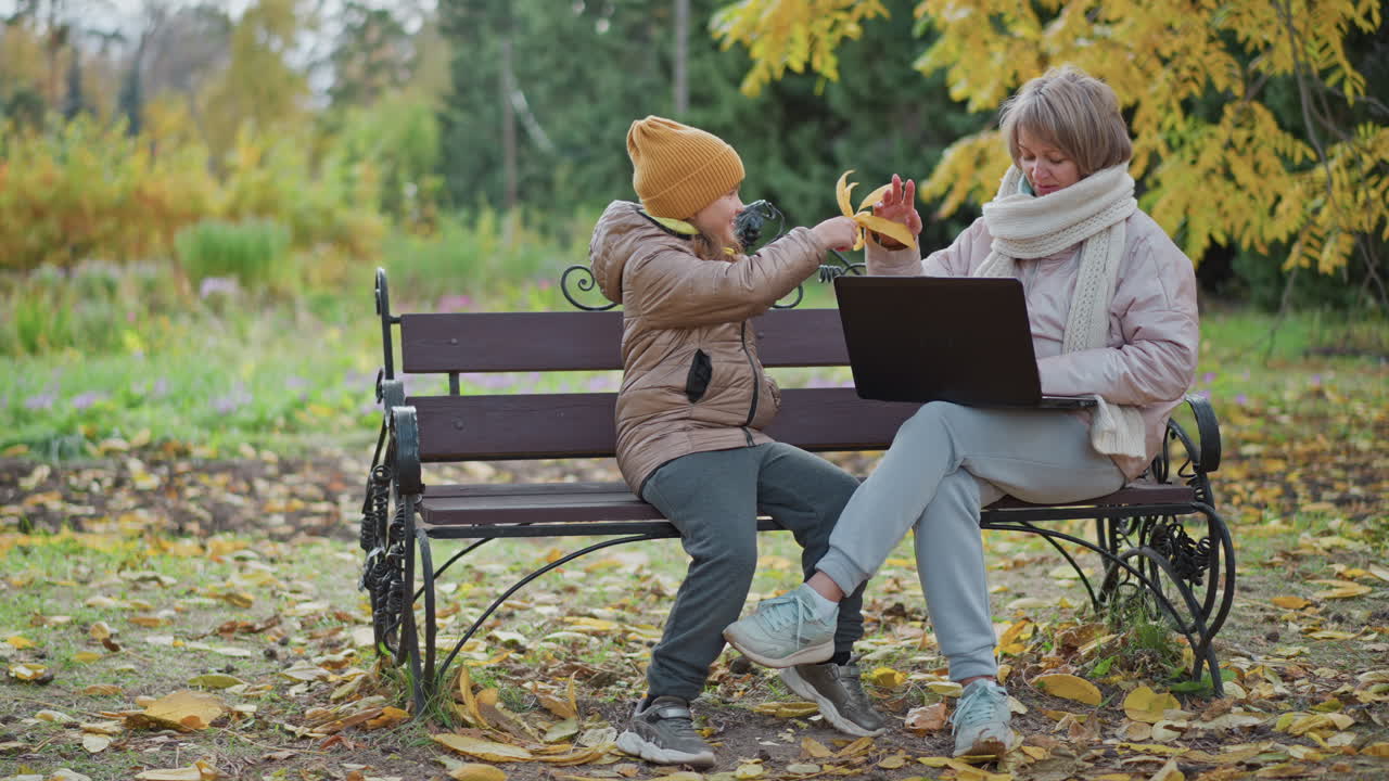 little girl rushing across ornate park bench waving bright yellow leaf excitedly to mum focused on laptop work outdoors amid scattered autumn leaves, warm scarf coat and cozy seasonal foliage