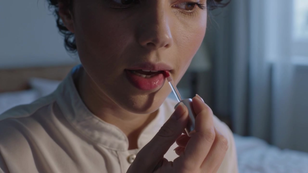 Frau in Oberteil mit Kragen trägt Lipgloss mit Applikator auf die Lippen auf, im Schlafzimmer, bereitet sich vor.