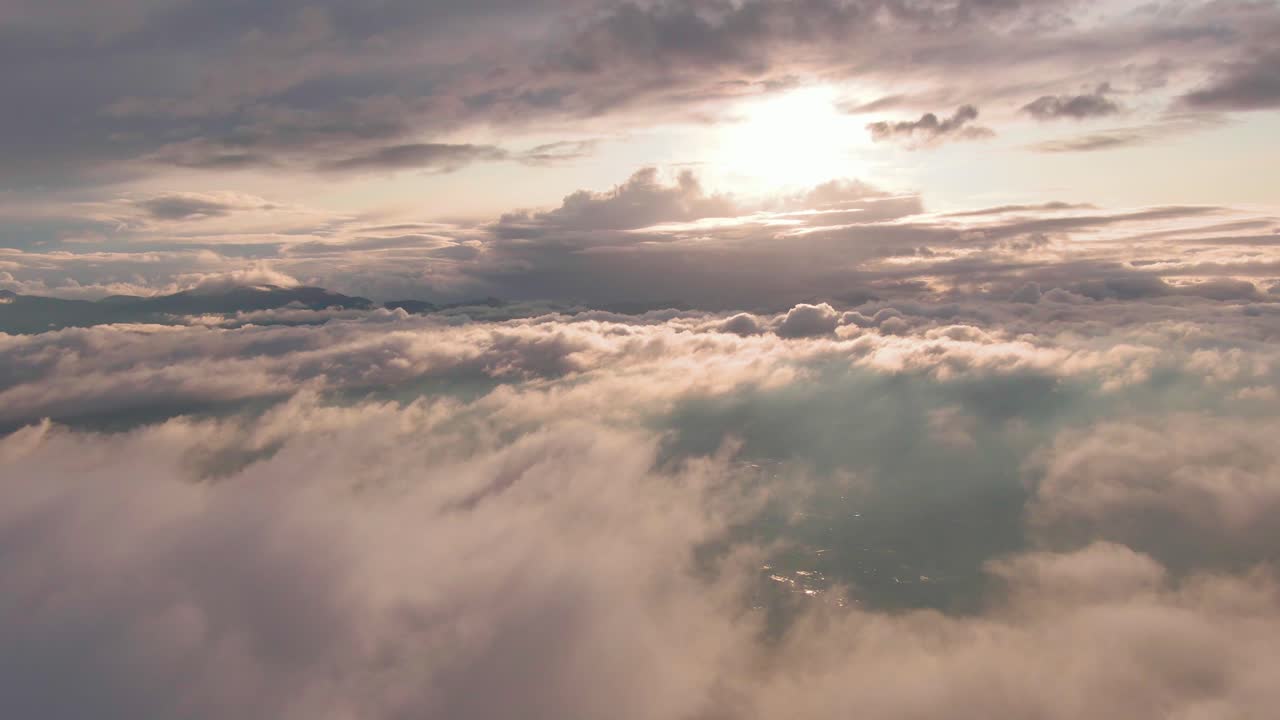 Volo panoramico sopra una coltre di nuvole soffici con un sole bianco e brillante e una catena montuosa all'orizzonte, vista aerea.