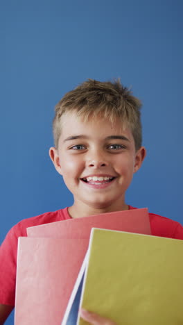 vertical video of happy caucasian boy holding books on blue background, copy space, slow motion