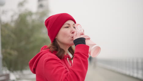 lady in red beanie and hoodie sipping water outdoors using pink bottle, against blurred background with greenery and iron rail, in serene winter setting