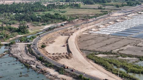 This elevated shot shows a winding road under construction, bordering water-logged fields and an expansive grid of salt pans, capturing the contrast between nature and industry