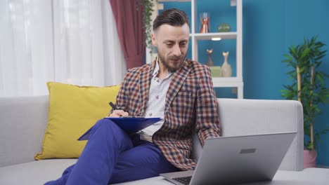 Happy-businessman-sitting-on-sofa-at-home-working-on-laptop-and-taking-notes.