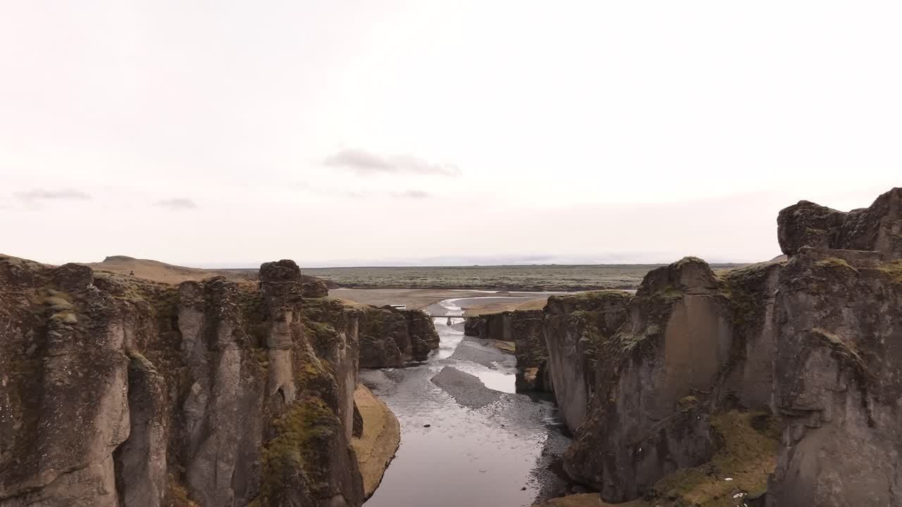 View from within Fjaðrárgljúfur Canyon, Iceland. The river flows between steep, mossy cliffs under an overcast sky. The bridge is visible in the distance.