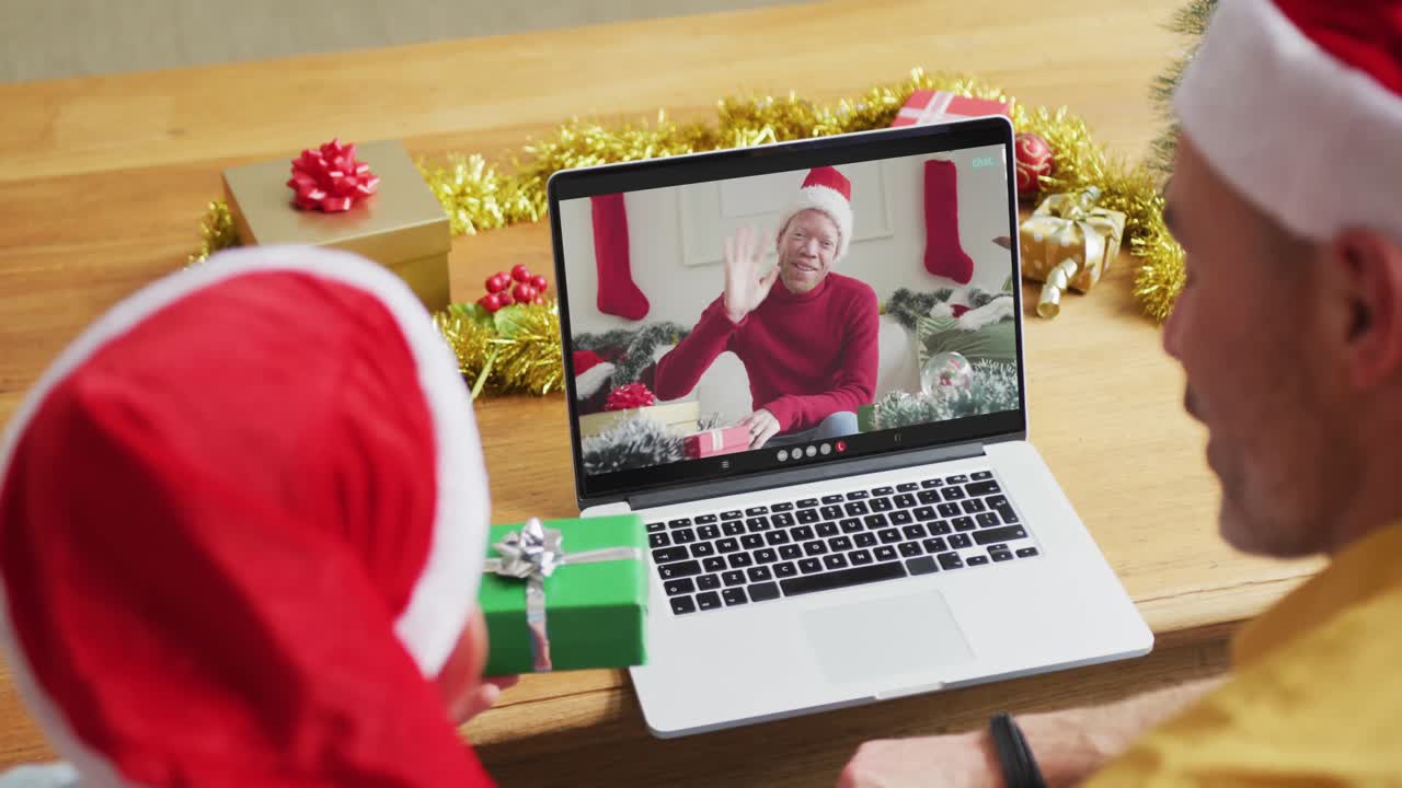 padre y hijo caucásicos con sombreros de santa usando una computadora portátil para una videollamada de navidad con un hombre en la pantalla