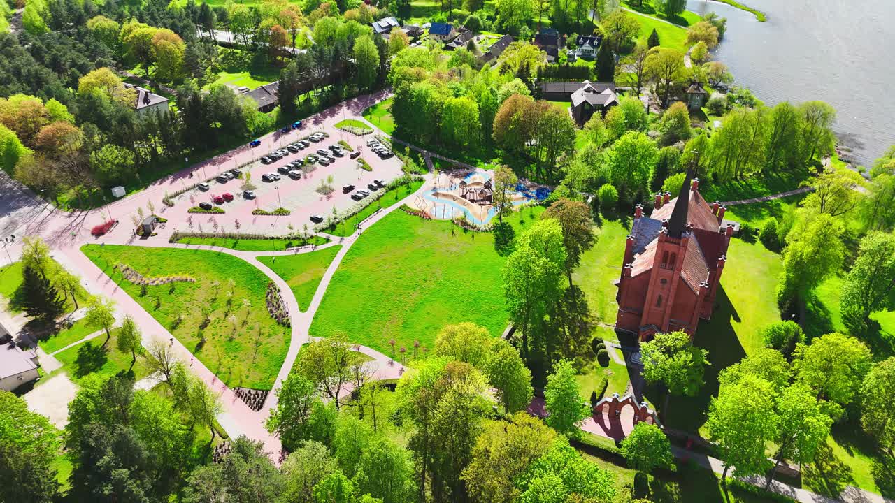 Aerial view of a historic church, playground, and parking lot within a lush riverside park. Bright spring foliage and well-kept pathways highlight urban greenery and recreation.