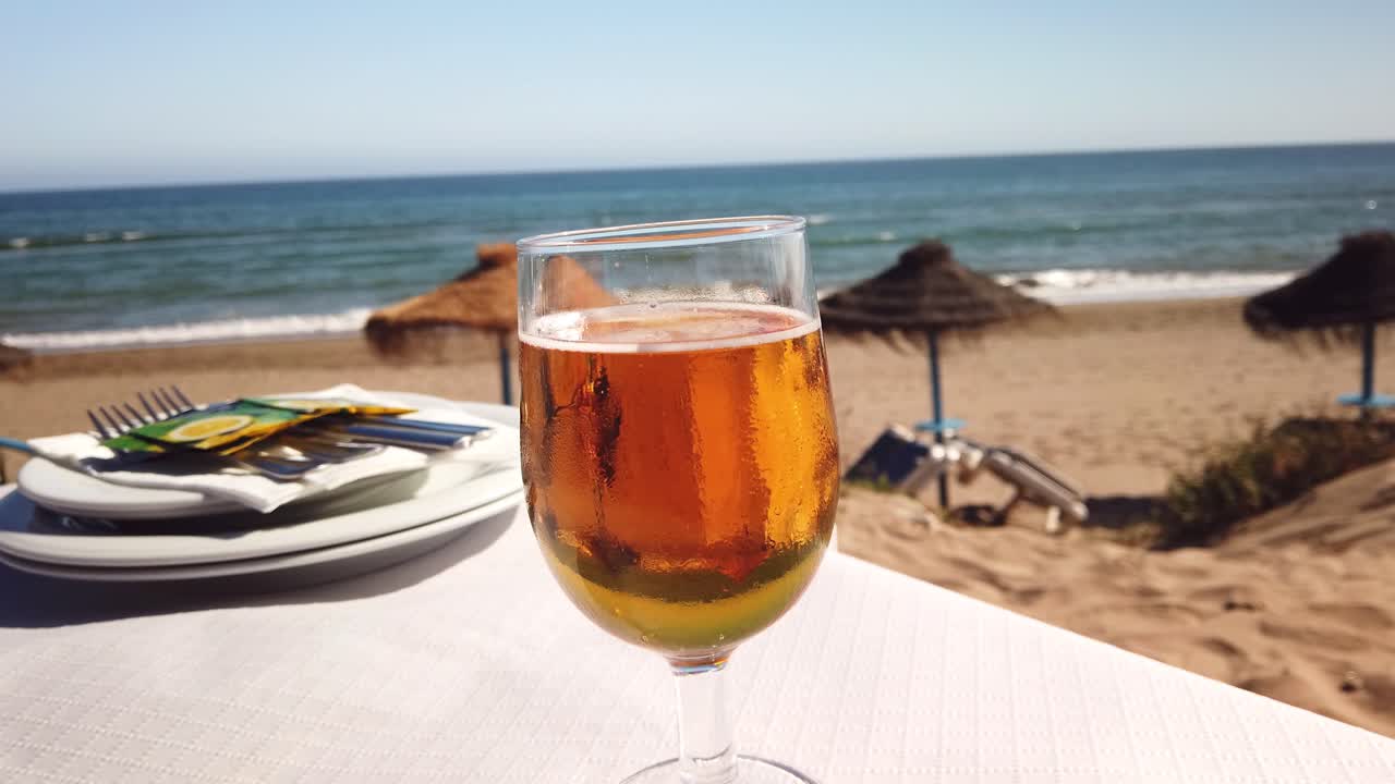 Bicchiere di birra fredda e rinfrescante su un tavolo con vista mare e una spiaggia spagnola a Marbella, filmato in 4K.