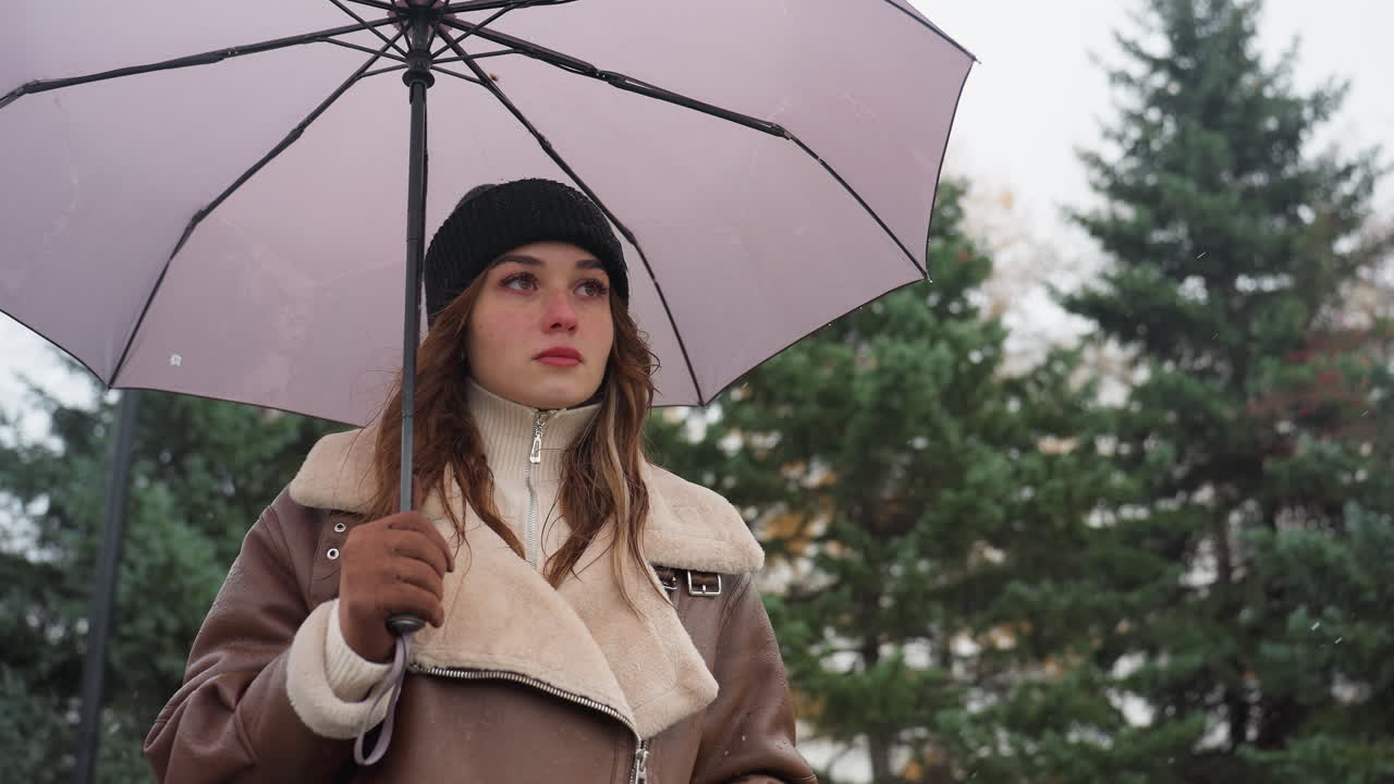Portrait shot of young woman holding umbrella walking calmly in black knit cap, brown shearling jacket, during cold overcast day with light snowfall, surrounded by greenery and cityscape