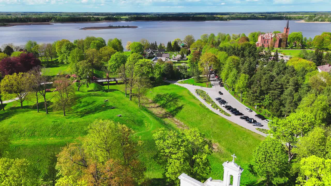 White church towers frame a lush riverside park where trees, footpaths, and a winding road lead to a distant red brick church across the spring landscape.